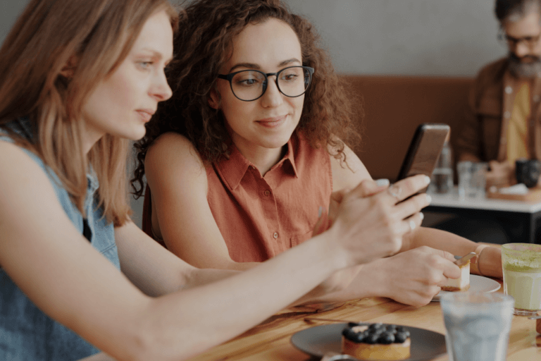 Dwie kobiety siedzące przy stoliku w kawiarni, wspólnie przeglądające zawartość smartfona.
